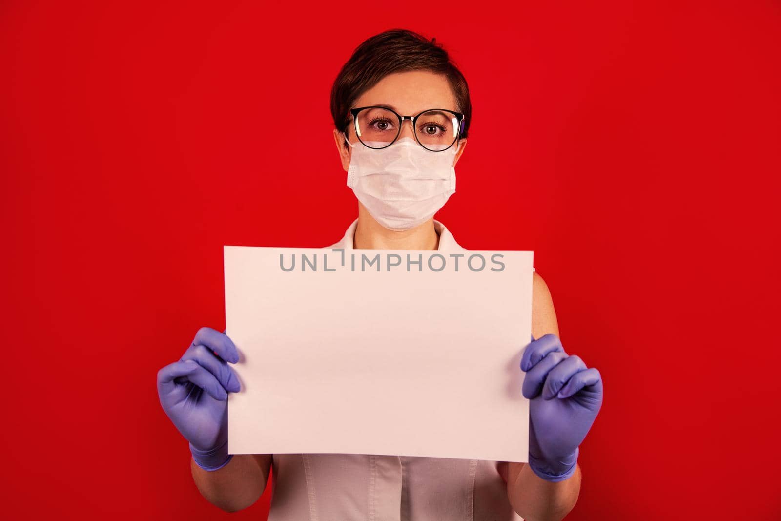 A nurse in a medical protective mask holds a blank white sheet on a red background. The concept of protection against viruses, influenza and coronavirus COVID-19. Health care and surgical concept.