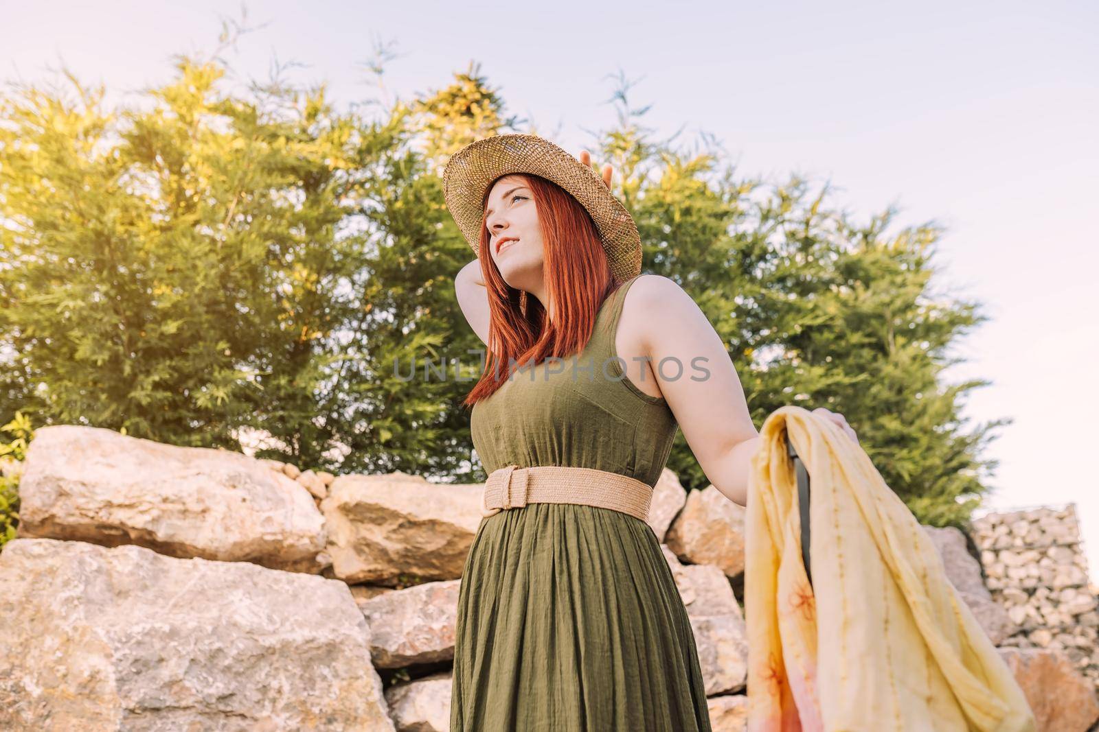 adventurous young girl, relaxing on her summer holiday. woman on a trip to the beach strolling along the coast. bathed in sunlight. woman in green summer dress, straw hat, outdoors with natural light and plants in background.