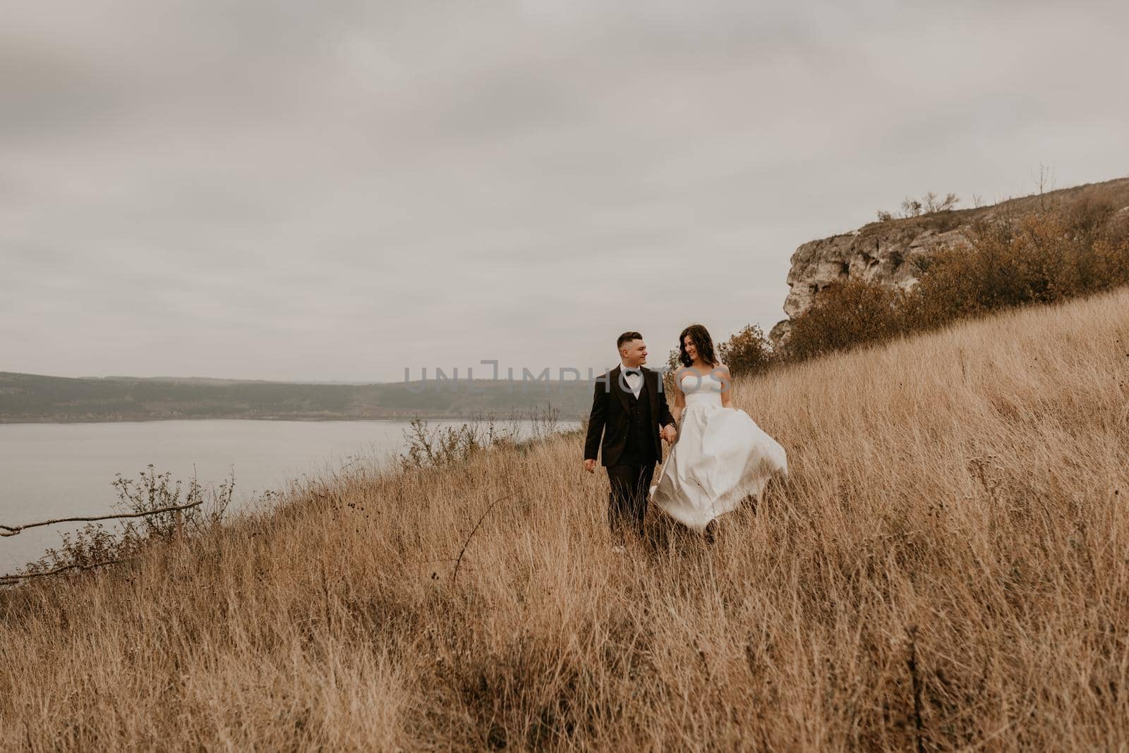 couple wedding newlyweds in walk hug kissing on tall grass on mountain above the river by AndriiDrachuk