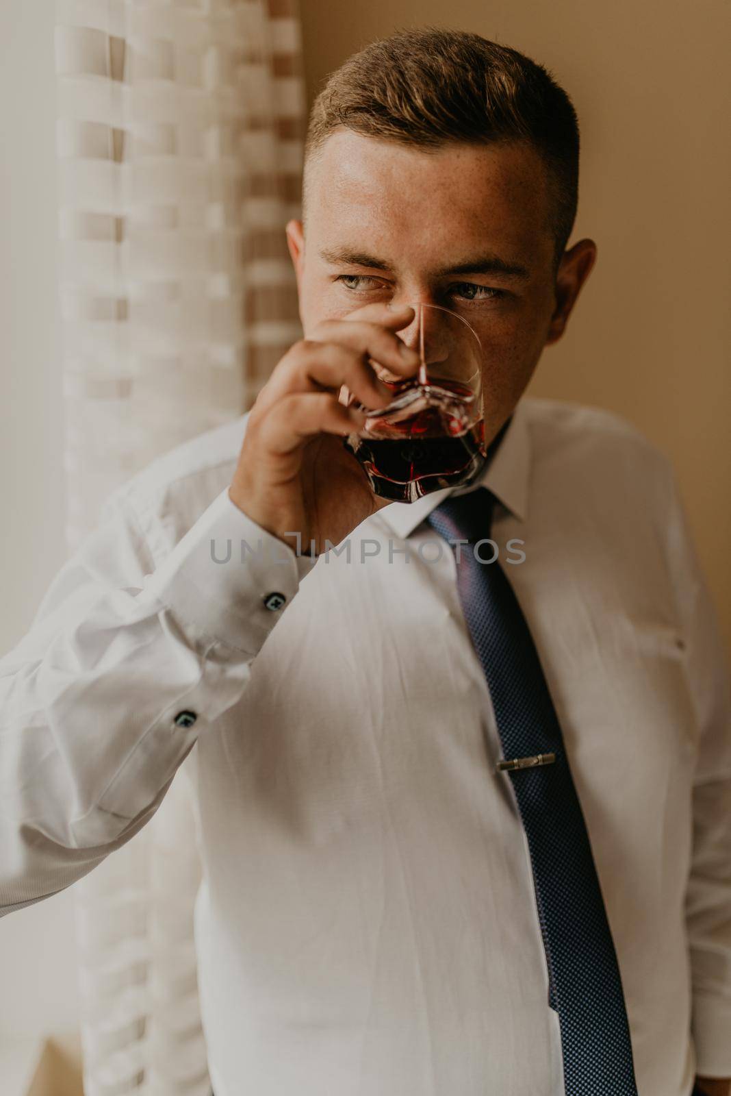 blonde European Caucasian young groom in shirt business suit with tie prepare for wedding. preparing for important event meeting indoors worries. a man holding a glass of whiskey in his hands drinking