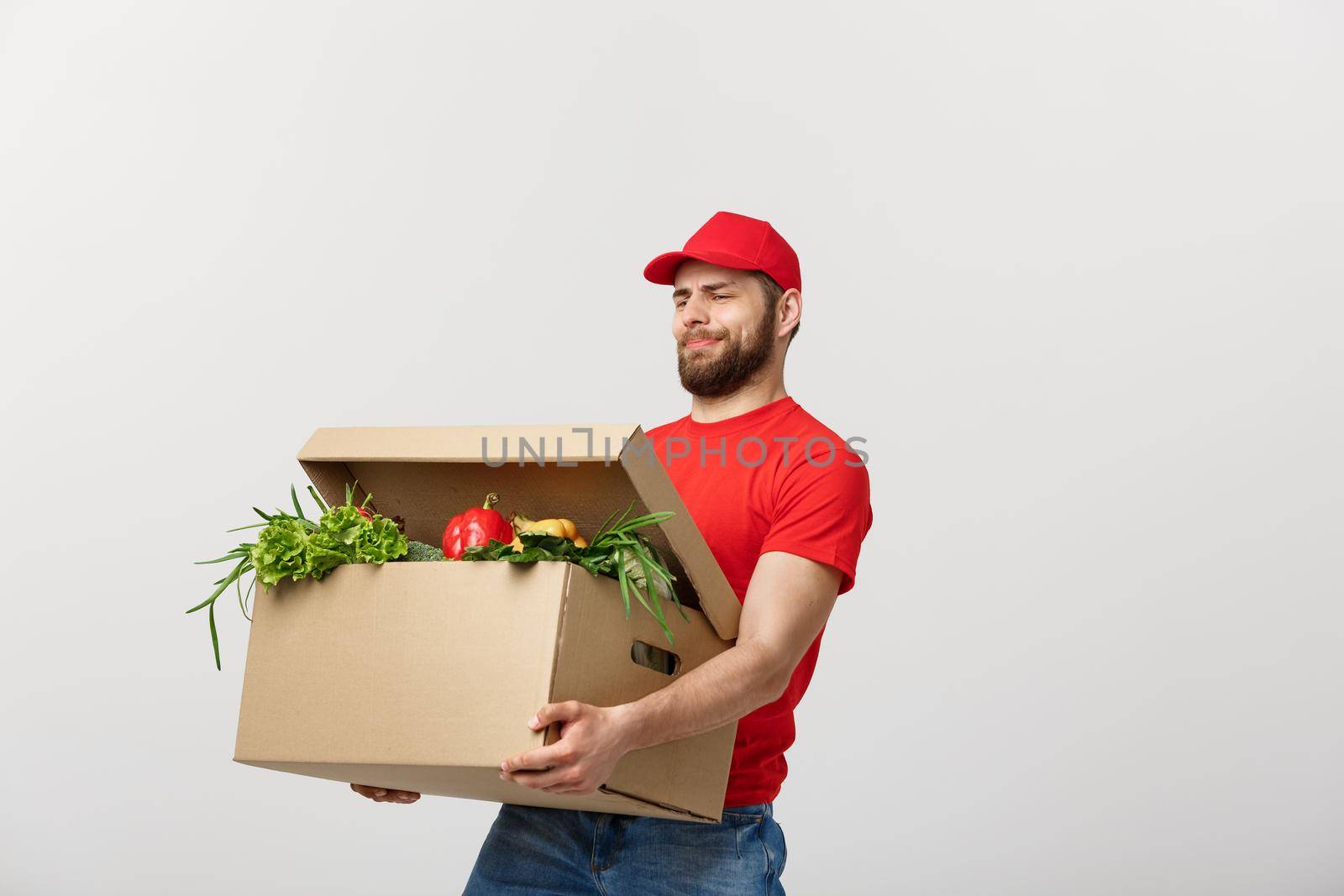 Delivery Concept: Handsome delivery man is holding a heavy grocery box isolated over grey background