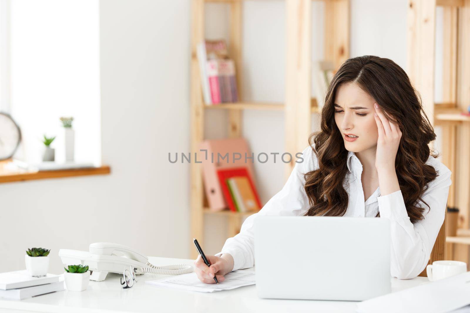 Tired beautiful Businesswoman holding hand on head while working on computer and some business documents in bright office.