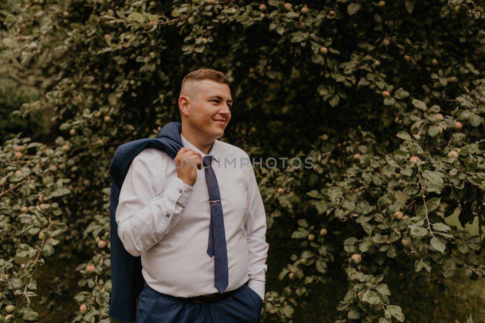 young blond man European curly-haired successful businessman groom in classic clothes white shirt and tie stands on street holding jacket thrown over his back shoulder in nature green leafy wall