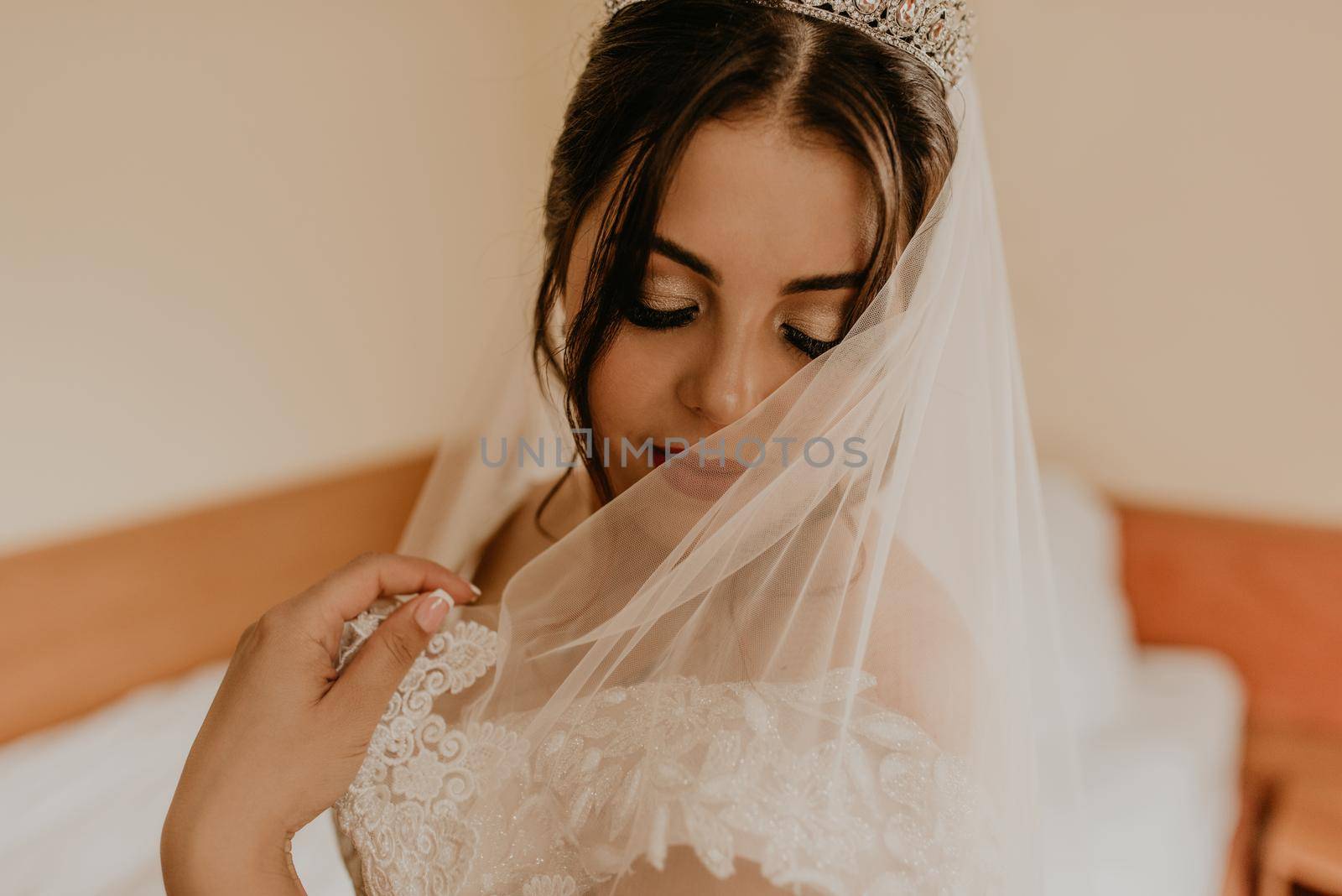 European Caucasian young black-haired woman bride in white wedding dress with long veil and tiara on head. girl bedroom in beige colors morning gatherings of preparing bride for holiday party wedding