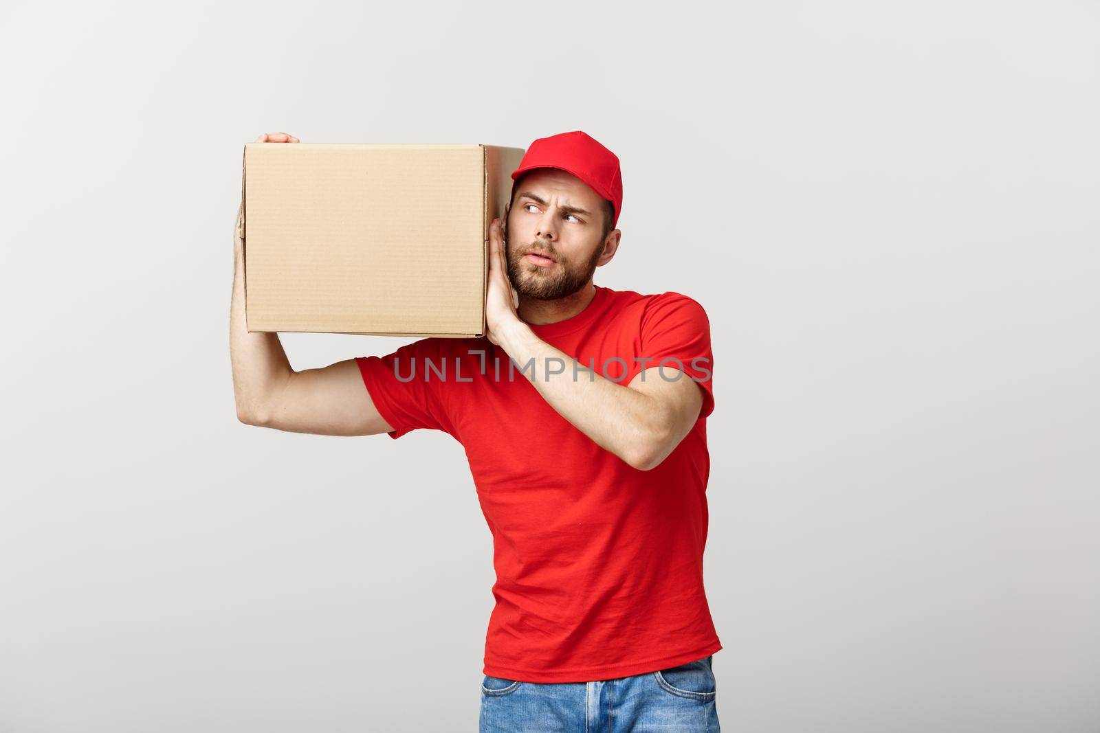 Delivery Concept - Portrait of Curious caucasian handsome delivery man listen inside a box package. Isolated on Grey studio Background. Copy Space