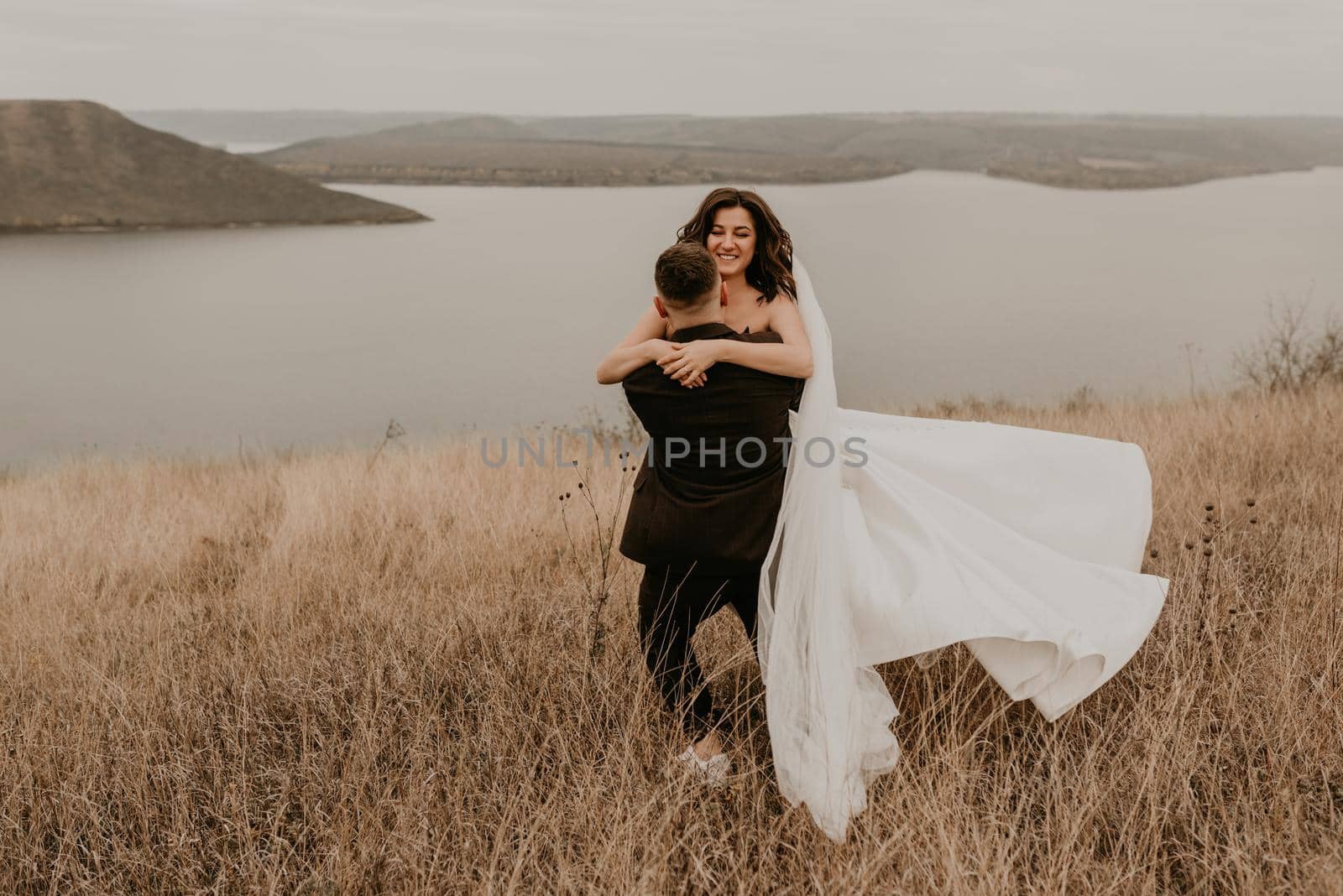 loving couple wedding newlyweds in dress veil sports shoes and suit hug kissing whirl on tall grass in field on mountain above river by AndriiDrachuk