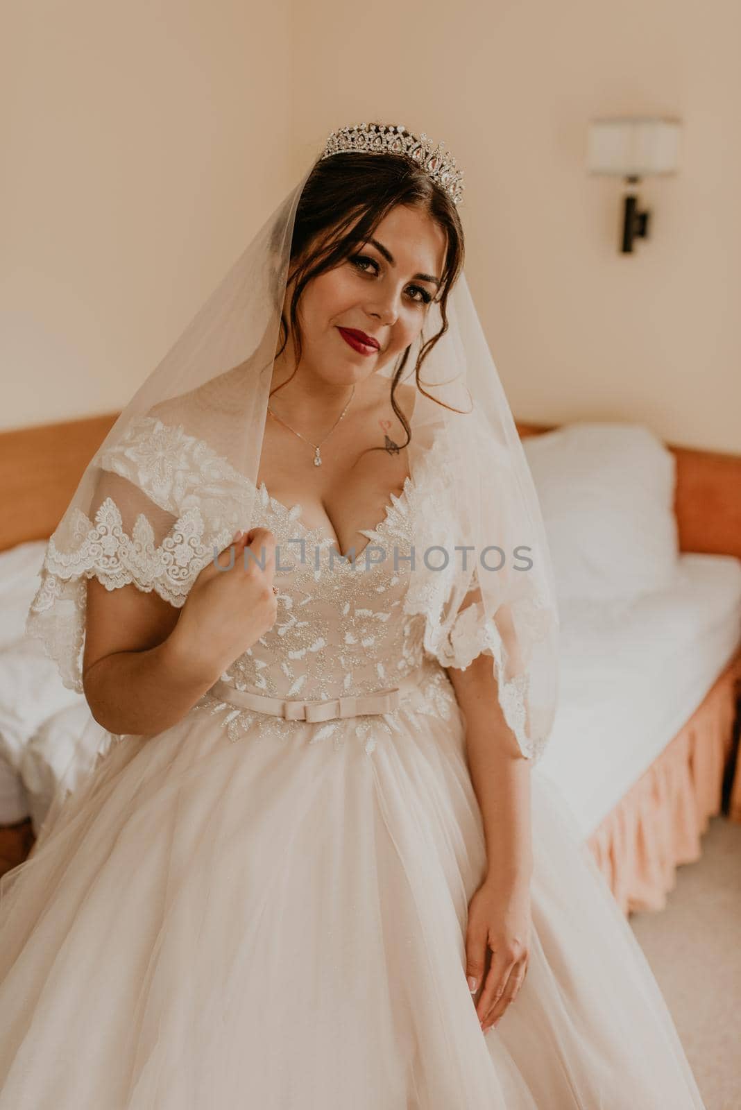 European Caucasian young black-haired woman bride in white wedding dress with long veil and tiara on head. girl bedroom in beige colors morning gatherings of preparing bride for holiday party wedding