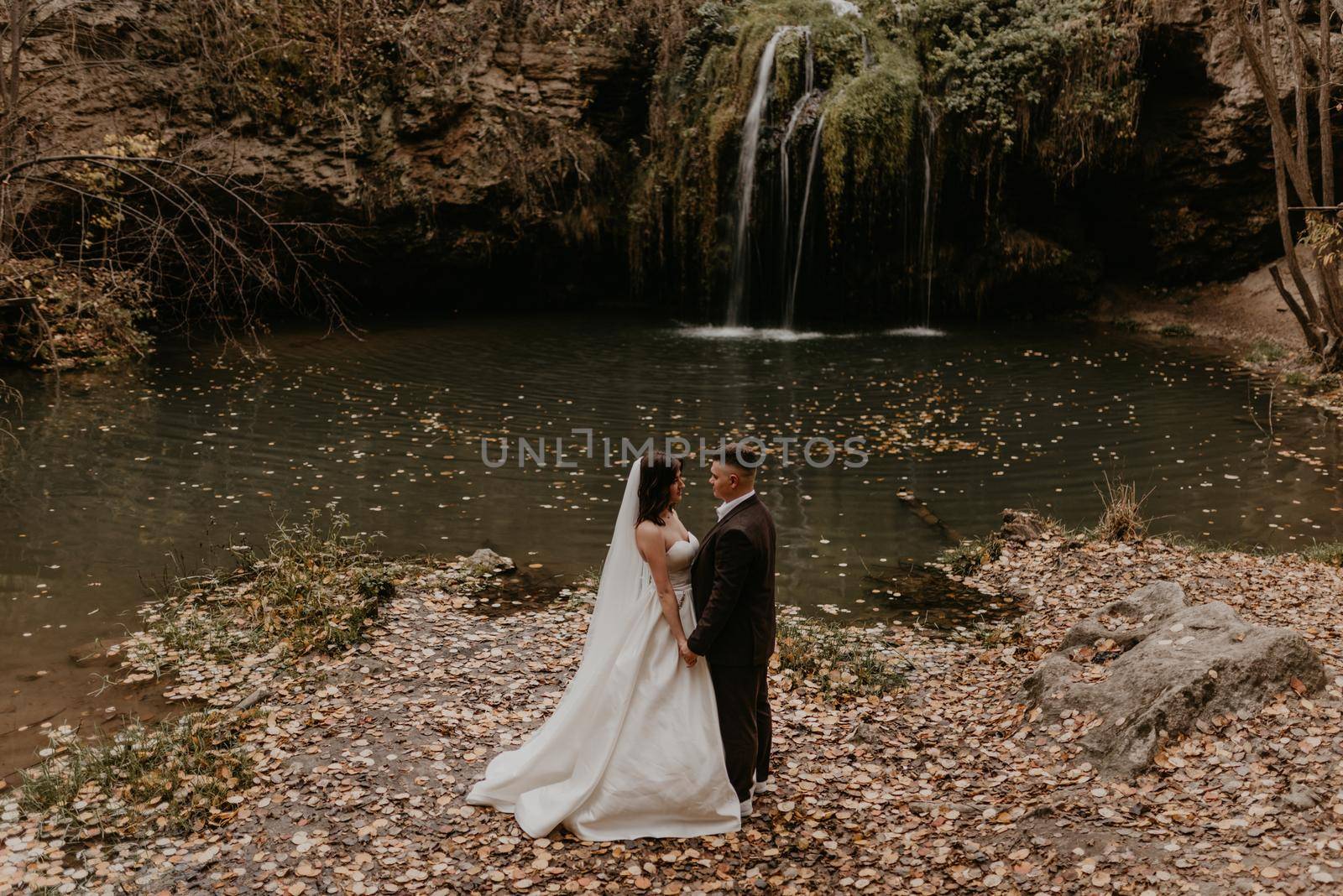 bride hugs groom near small pond lake river waterfall Burbun by AndriiDrachuk