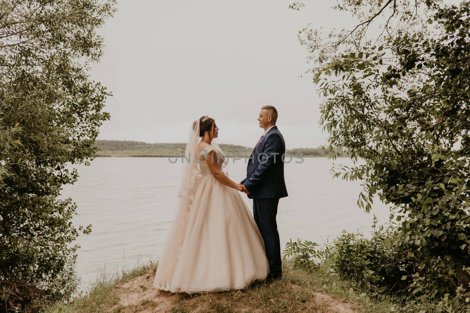 blonde European Caucasian young man groom in blue suit and black-haired woman bride in white wedding dress with long veil and tiara on head. Newlyweds hold hands smile kiss and look at each other near river