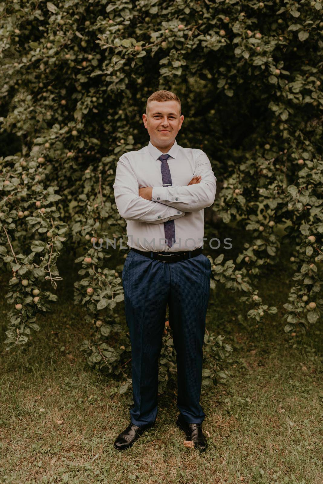 young blond man European curly-haired successful businessman groom in classic clothes white shirt and tie stands on street with his hands clasped together in nature on background of a green leafy wall