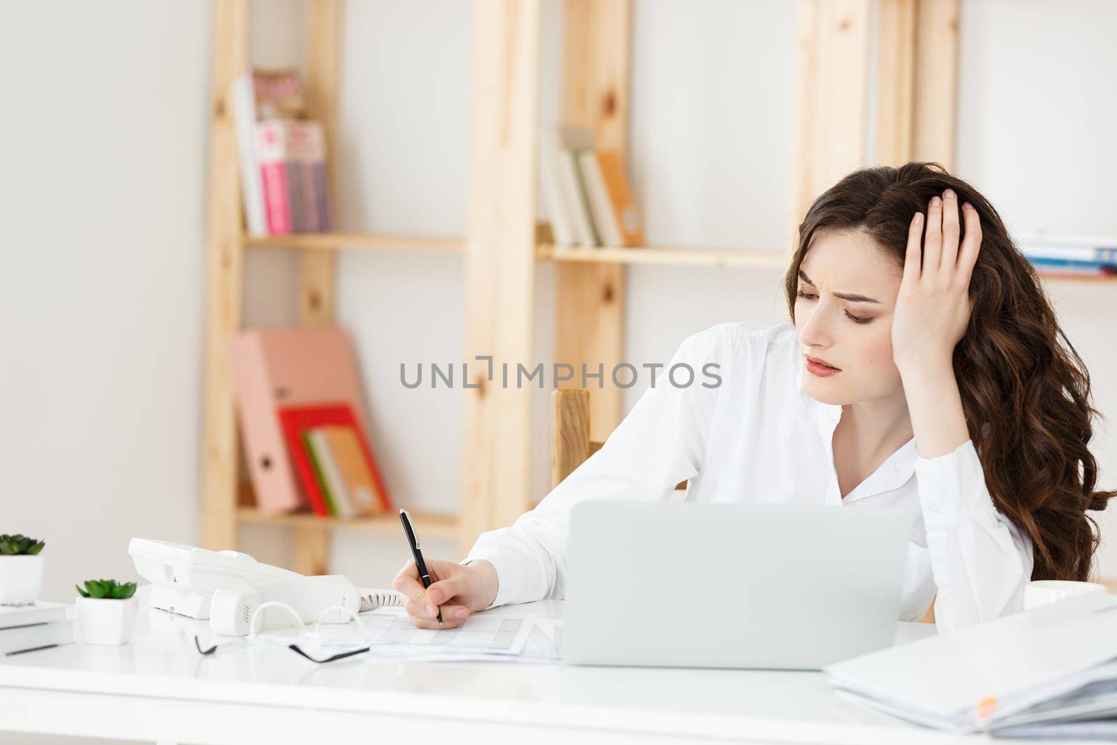 Tired beautiful Businesswoman holding hand on head while working on computer and some business documents in bright office.