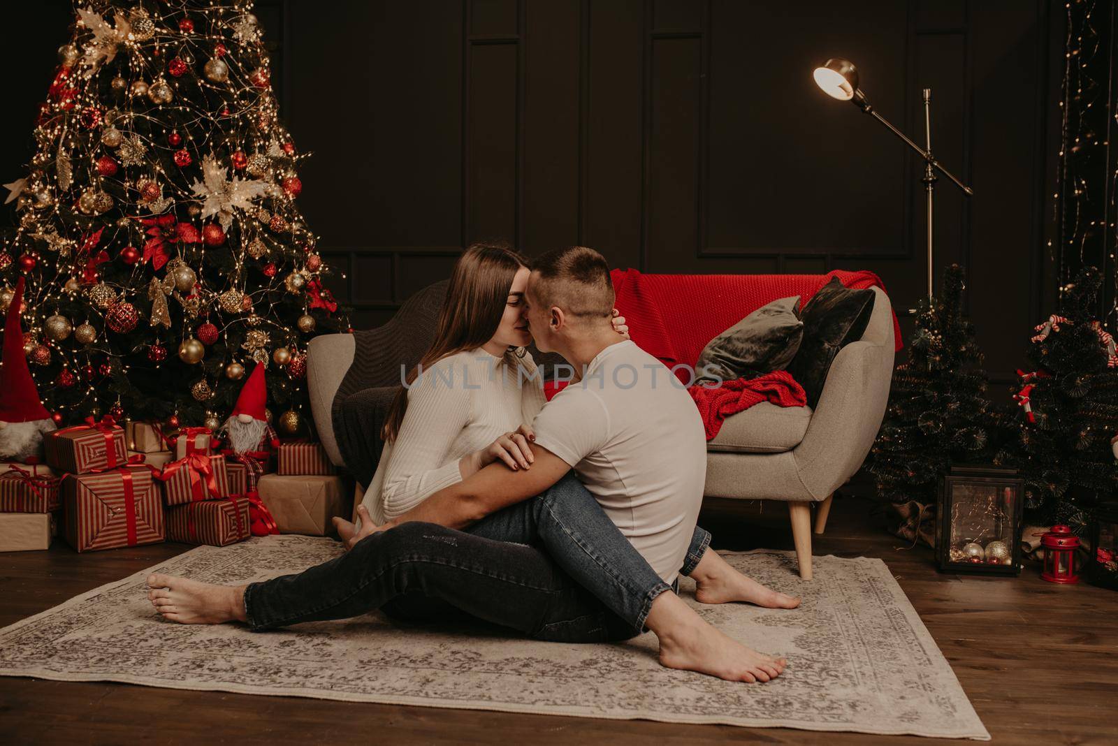 in love couple man and woman sitting on floor cross-legged embrace hugging and kiss Christmas tree. decorated house for New Year. Christmas morning. apartment interior. Valentine's Day celebration