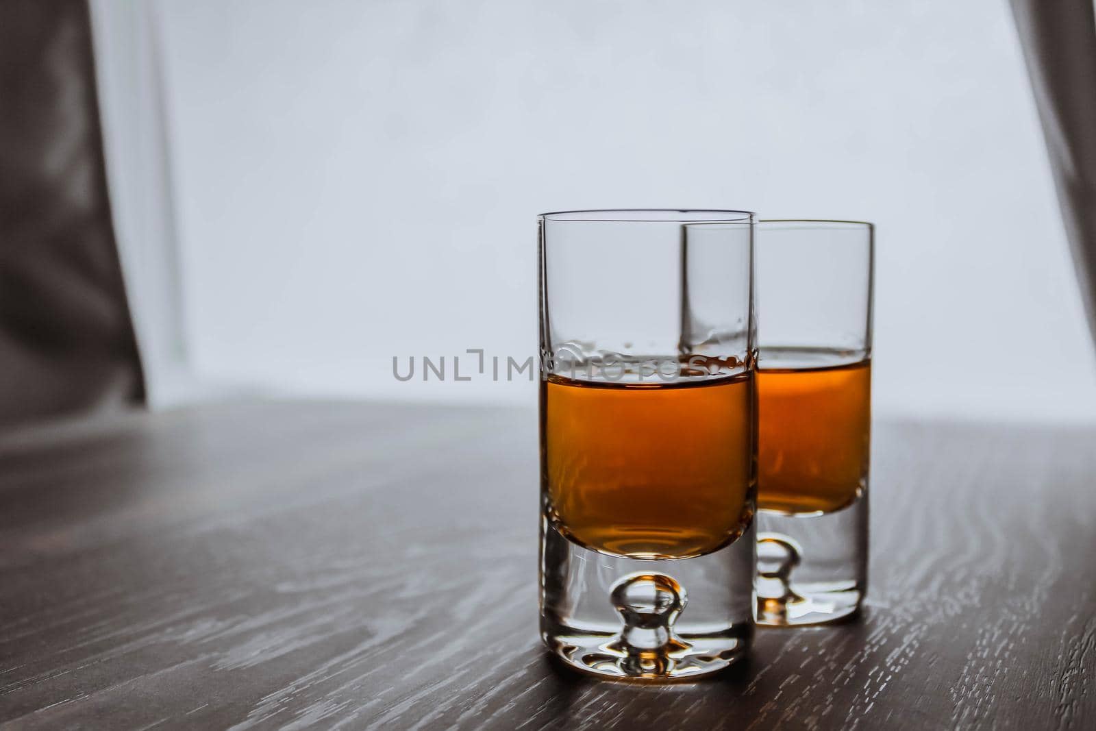 Two shot glasses with whiskey on a wooden background table next to window. Space for text.