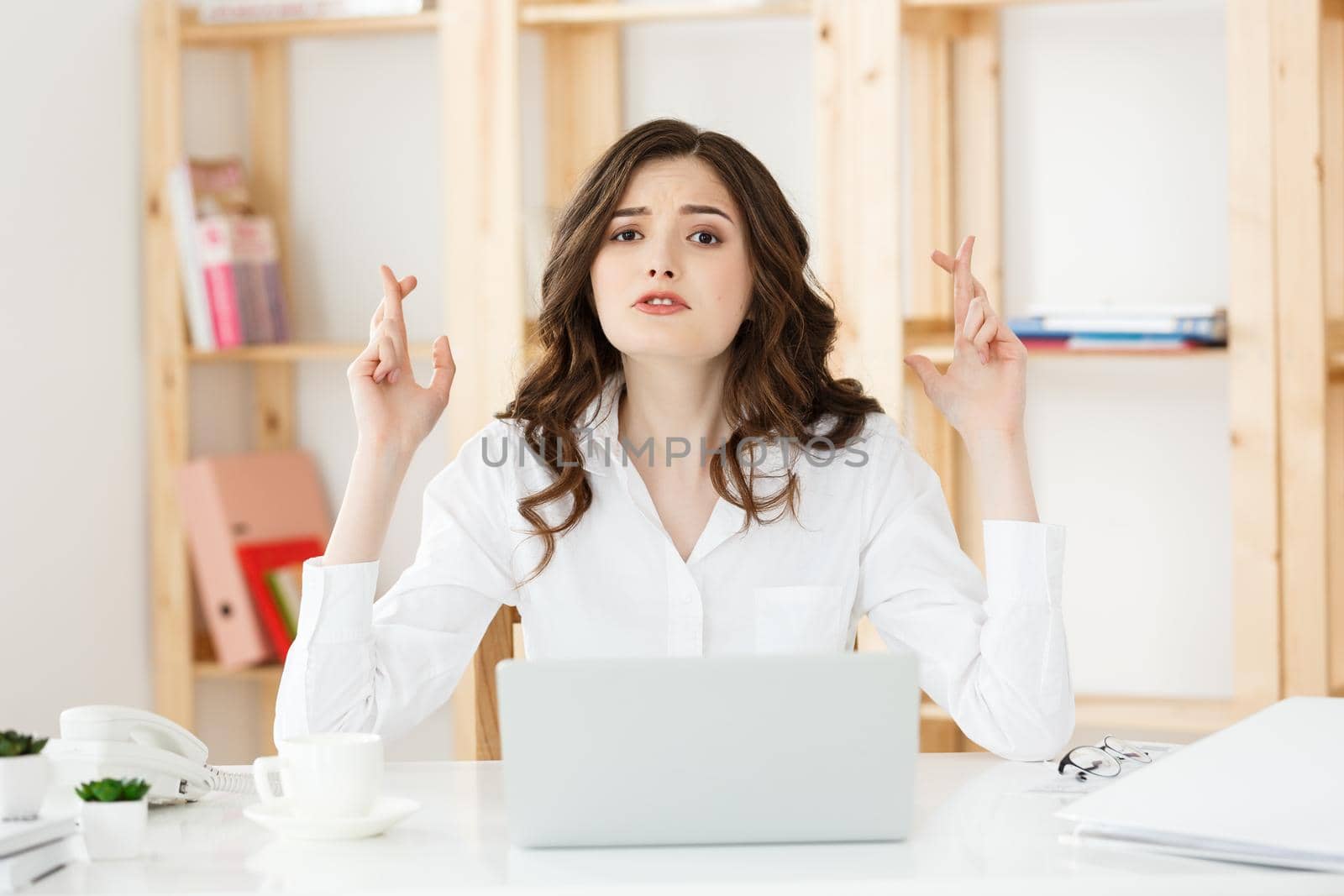 Portrait of happy young caucasian business woman sitting at the office desk and holding fingers crossed isolated over white background.