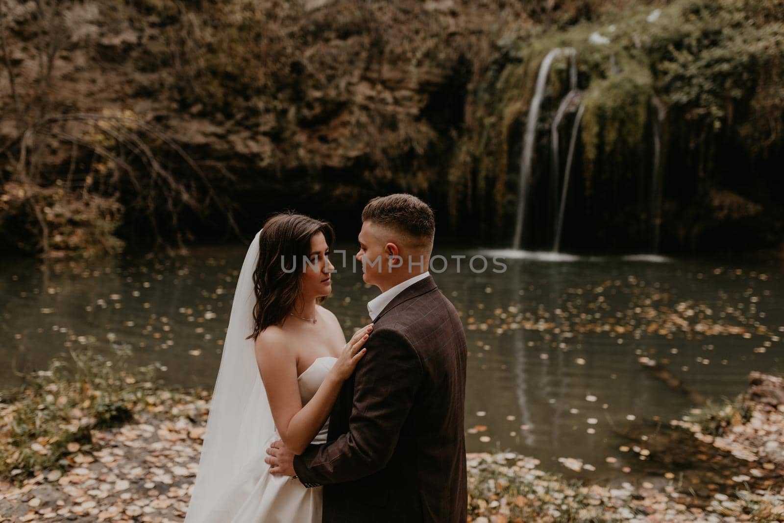 bride in white wedding dress and long veil hugs groom in a suit small pond lake river waterfall Burbun by AndriiDrachuk