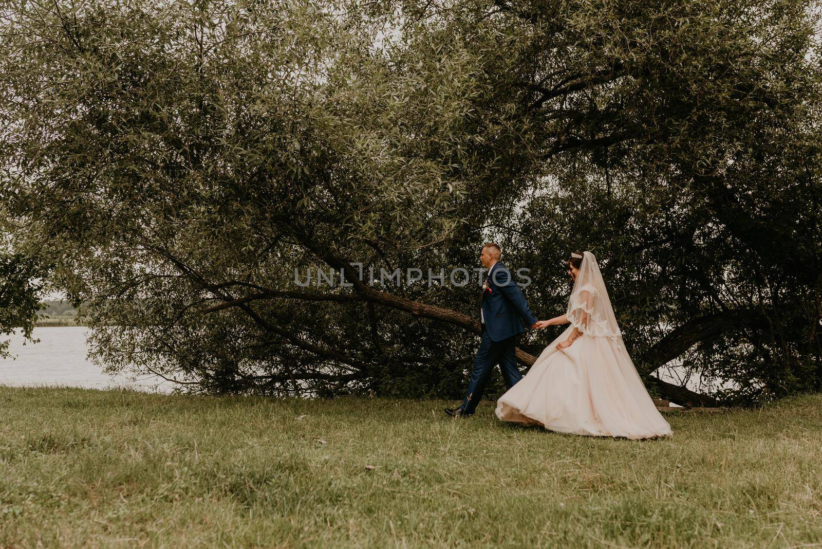 blonde European Caucasian young man groom in blue suit and black-haired woman bride in white wedding dress long veil and tiara on head. Newlyweds walk holding hands along river in summer near trees