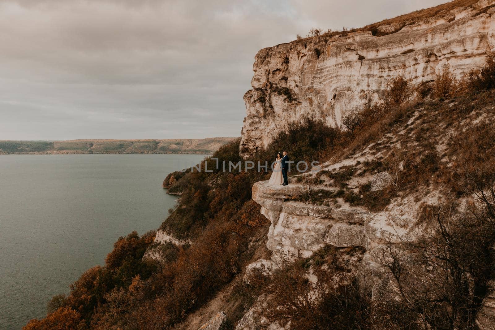 groom in suit and the bride in a dress stand on a stone cliff above the river. Mountain near lake. Aerial view from drone. rock monastery in bakota