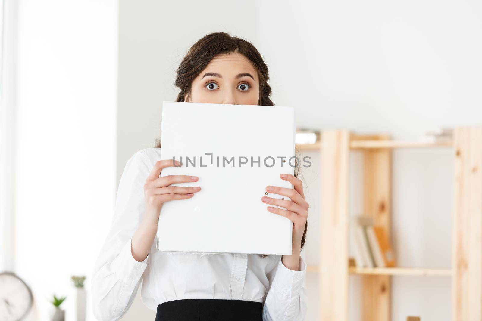 Young woman with surprised eyes peeking out from behind paper poster. Businesswoman holding big white banner in modern office