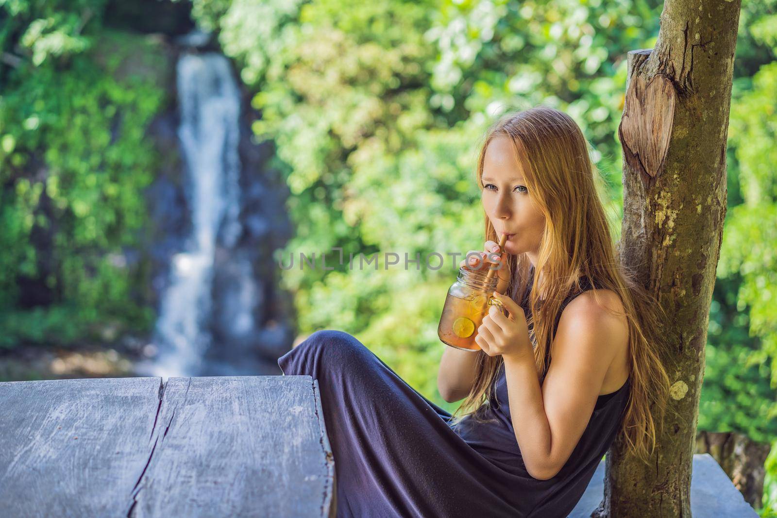 Closeup portrait image of a beautiful woman drinking ice tea with feeling happy in green nature and waterfall garden background.