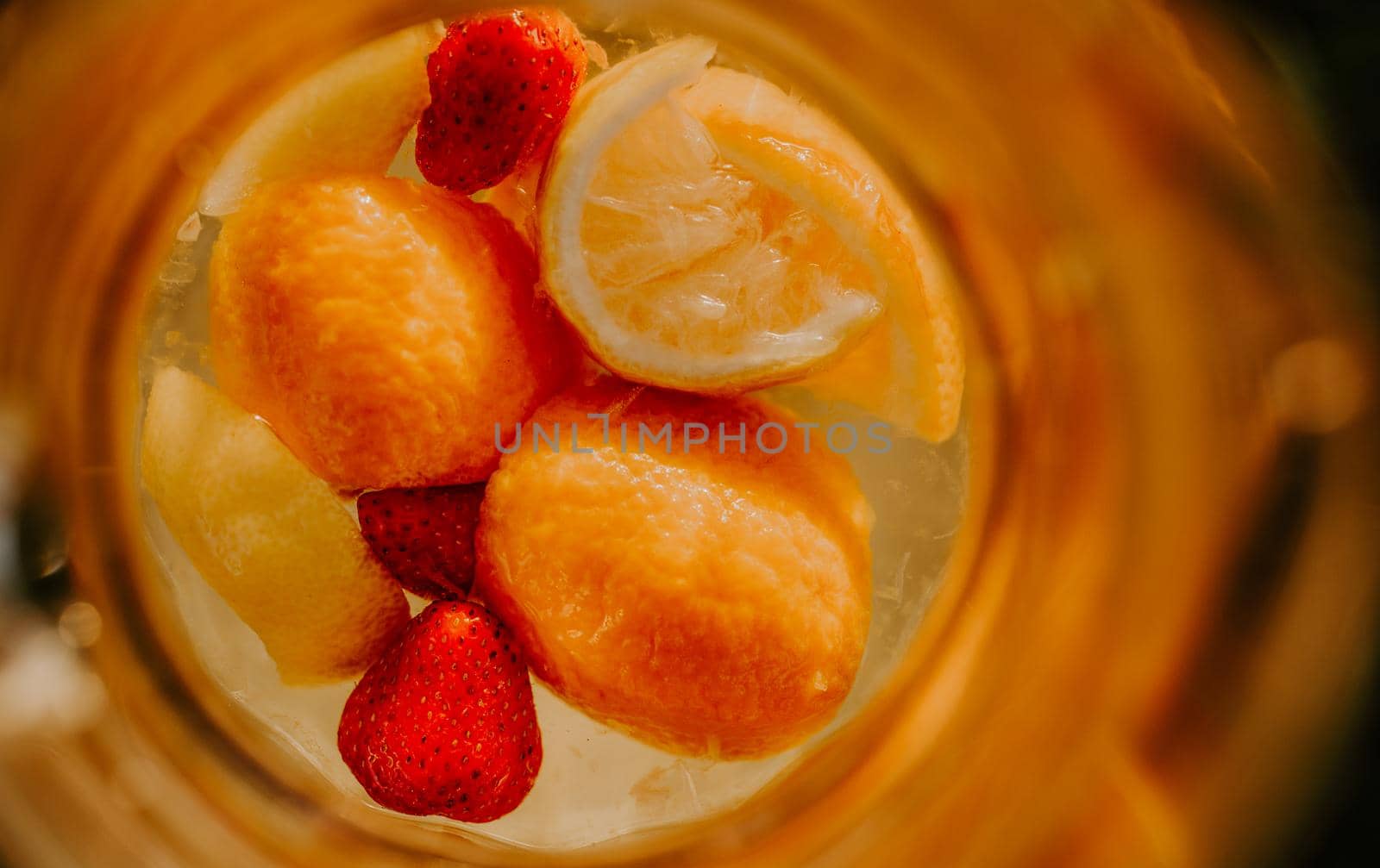 A glass jug with lemonade and fruit inside is standing on a wooden stump. at sunset on a background of green grass. strawberries, orange, lemon and mandarin.