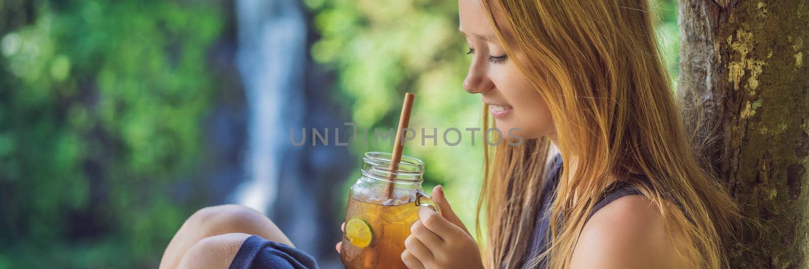 Closeup portrait image of a beautiful woman drinking ice tea with feeling happy in green nature and waterfall garden background. BANNER, LONG FORMAT