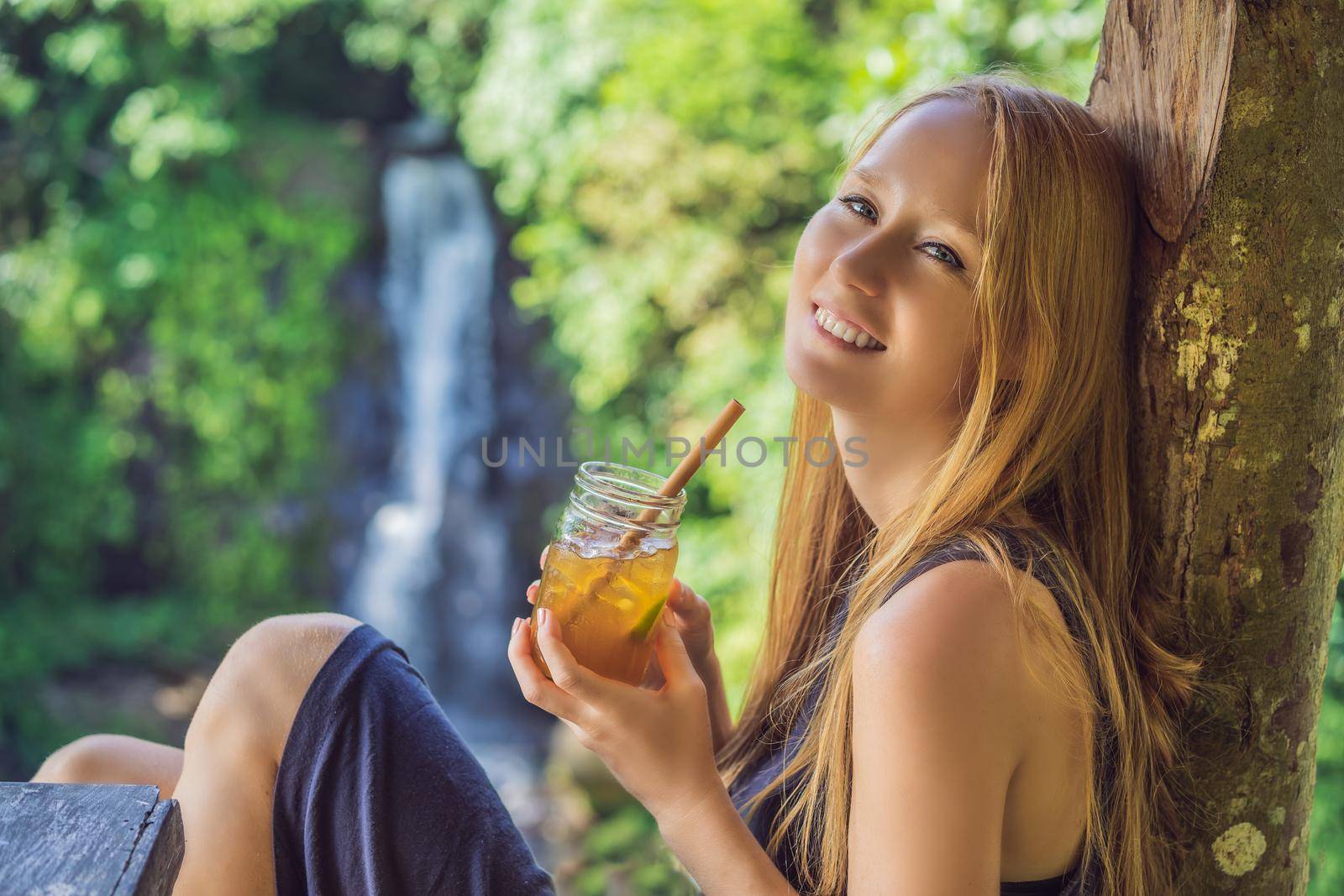 Closeup portrait image of a beautiful woman drinking ice tea with feeling happy in green nature and waterfall garden background.