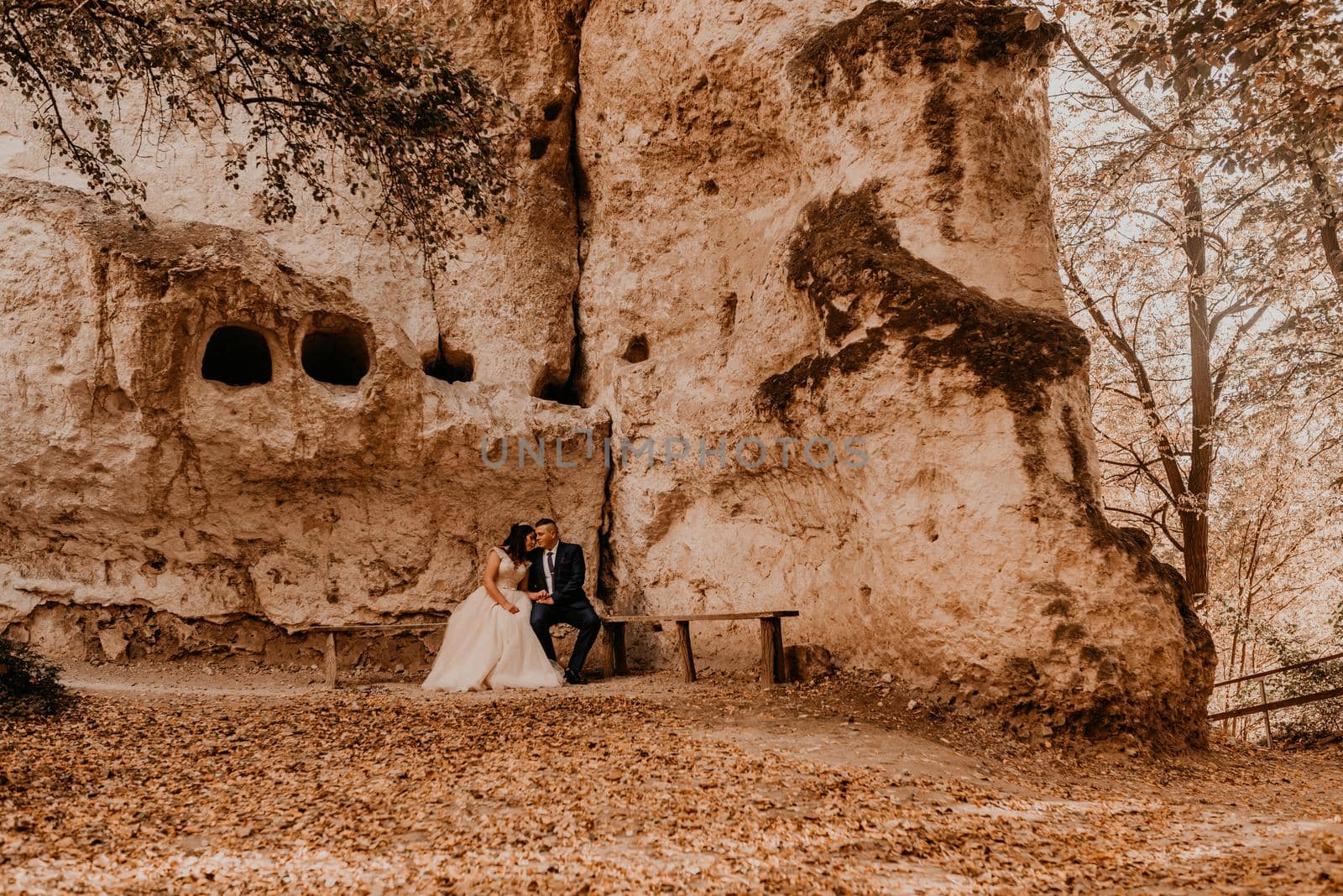 wedding couple man and woman sitting on a bench under rock monastery in bakota in autumn forest by AndriiDrachuk