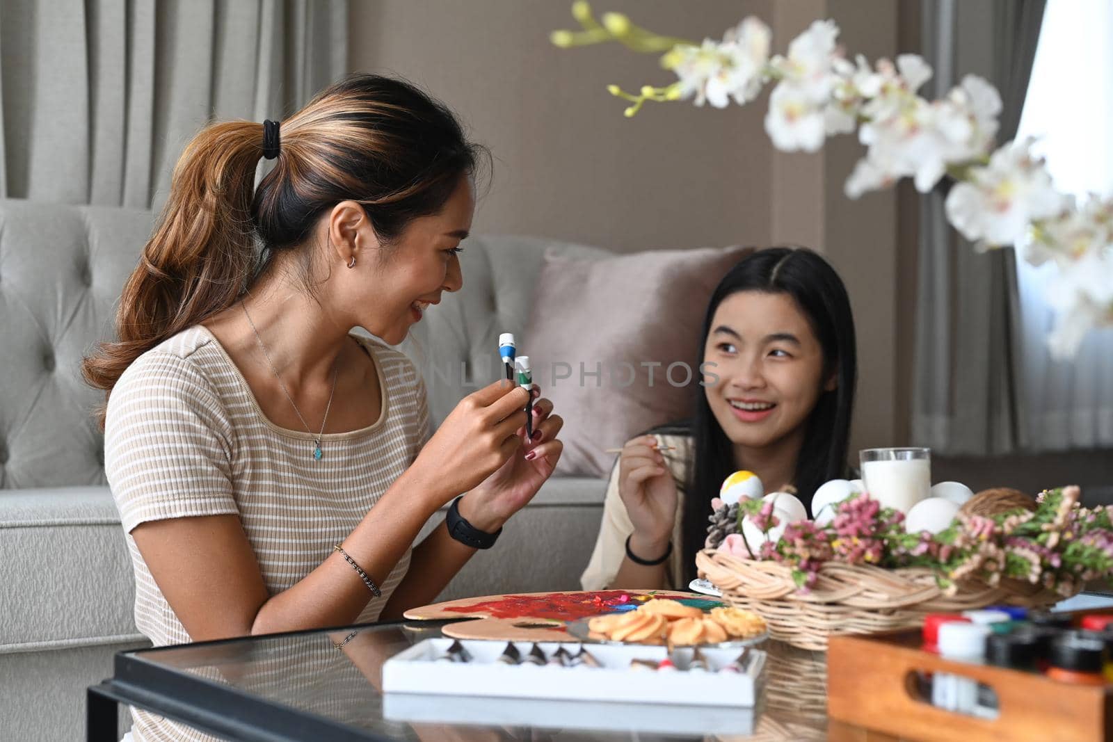 Happy girl and mother talking each other while painting Easter eggs together at home. Easter holidays and people concept.