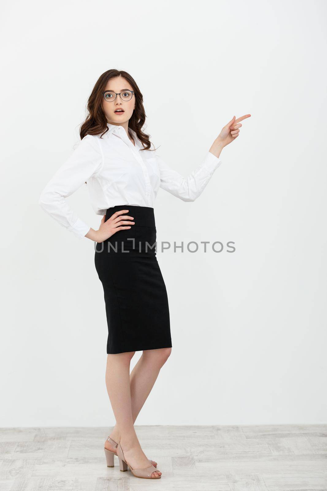 Full length portrait of a happy young businesswoman with eyeglasses standing and pointing finger away at copy space isolated over gray background.