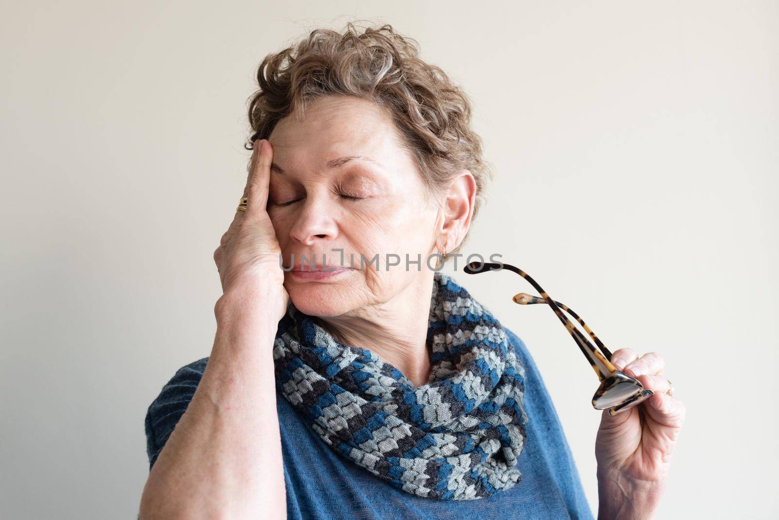 Head and shoulders view of older woman in blue clothing rubbing eyes and holding glasses (selective focus)