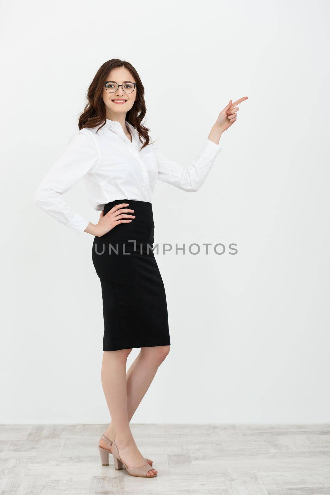 Full length portrait of a happy young businesswoman with eyeglasses standing and pointing finger away at copy space isolated over gray background.