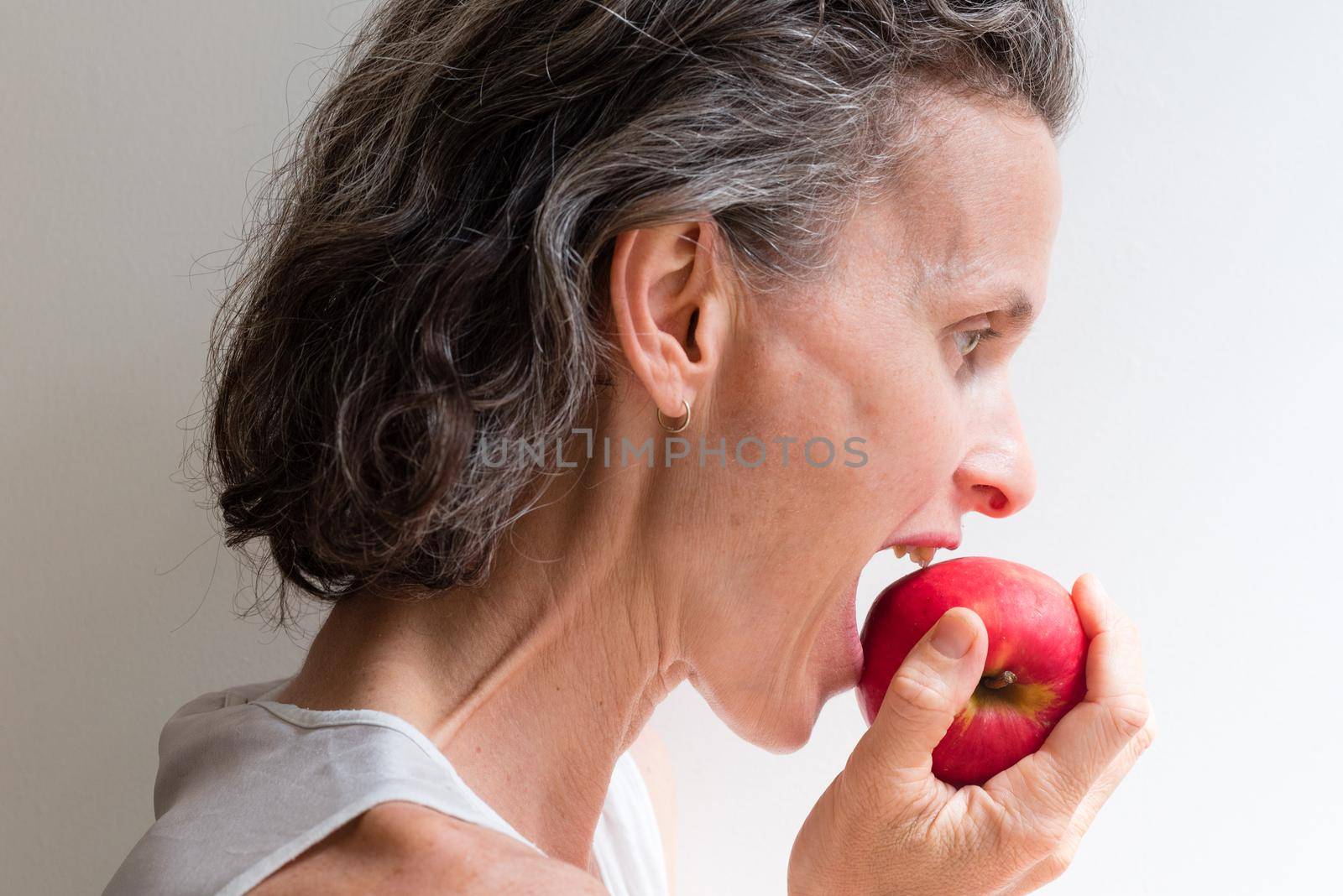 Profile of middle aged woman with grey hair biting into red apple (selective focus)