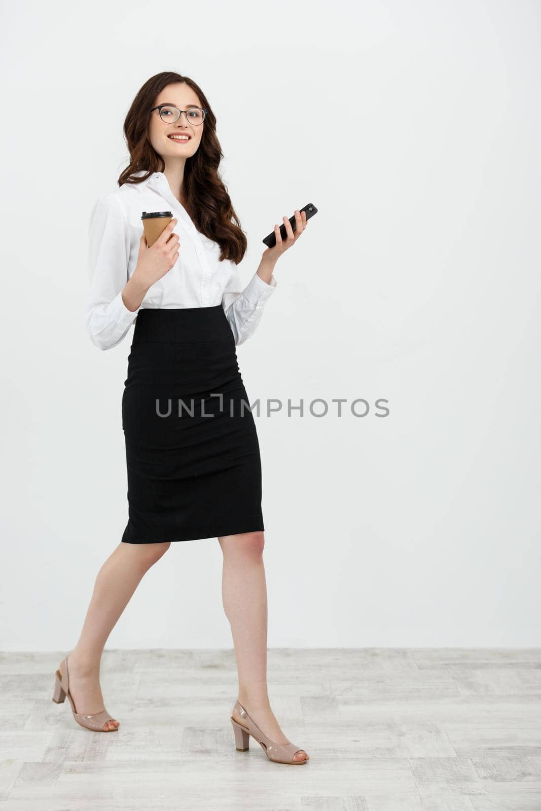 Full length Portrait of beautiful young business woman in formal wear walking and texting on cell phone with takeaway coffee in hand isolated over gray background.