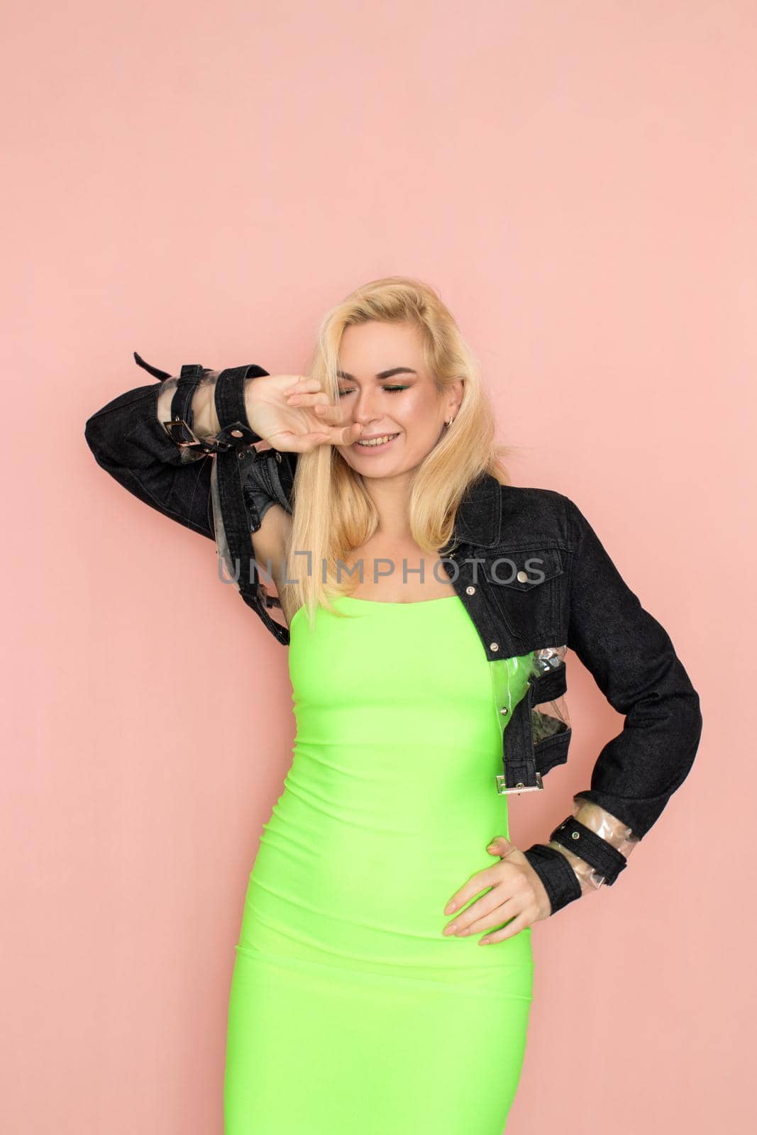 Stylish caucasian adult blonde woman wears fashionable jeans jacket and green dress, isolated over pink studio wall.