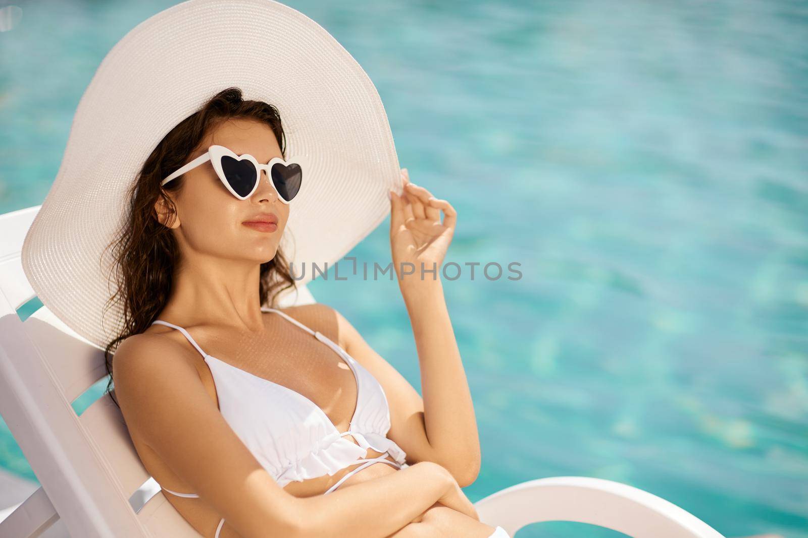 Enjoyed young girl in sunglasses with heart shapes, sunbathing unde sun, while sitting near pool at sunny day. Front view of calm beauty in wide hat, resting on lounger outdoor. Concept of relax.