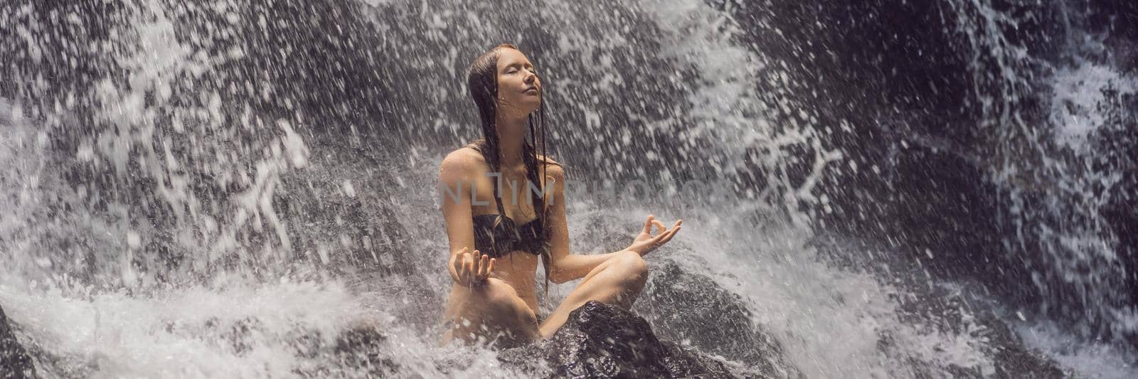 Wellness spa, vacation and yoga meditation concept. Young woman sitting in lotus position on the rock in tropical waterfall. BANNER, LONG FORMAT