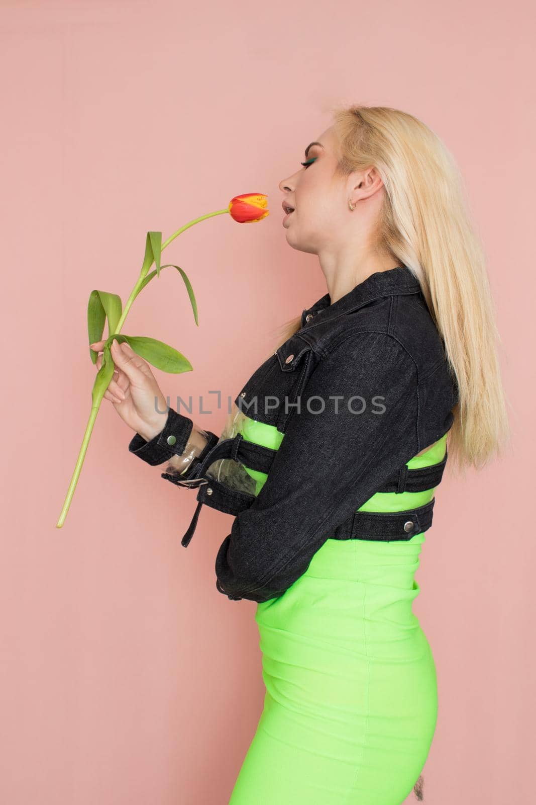 Young elegant woman in trendy jeans jacket. Blonde hair, green dress, isolated over pink background, studio shot. Fashion spring lookbook. Model woman with red tulips in her hands