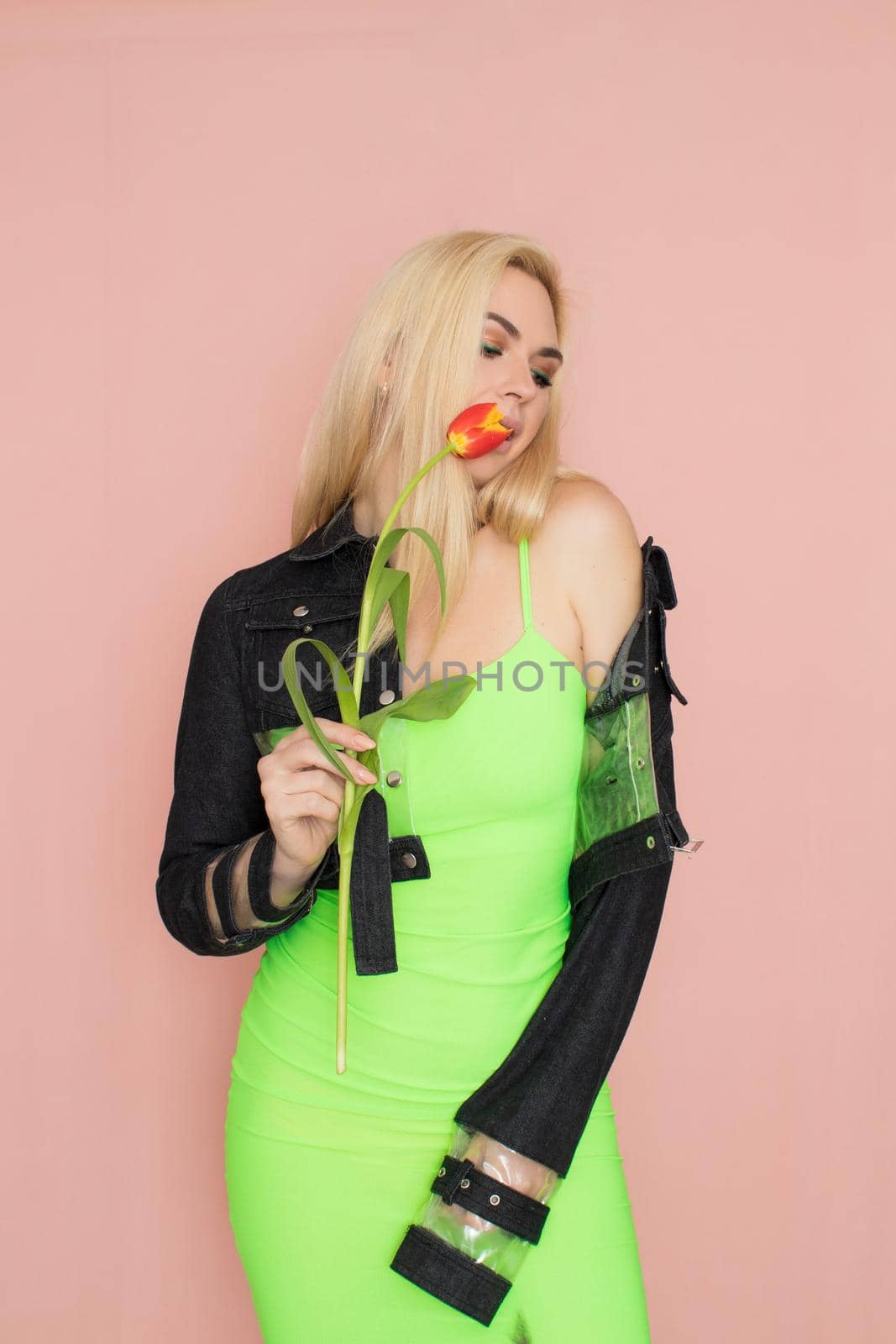 Young elegant woman in trendy jeans jacket. Blonde hair, green dress, isolated over pink background, studio shot. Fashion spring lookbook. Model woman with red tulips in her hands