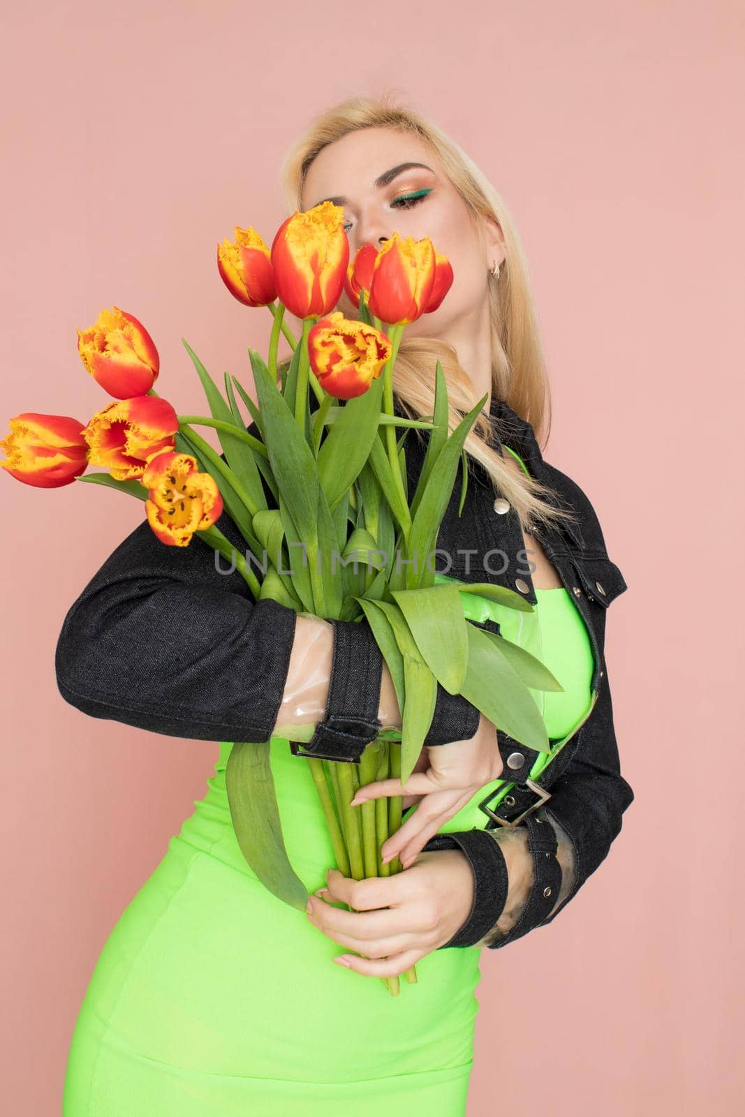 Young elegant woman in trendy jeans jacket. Blonde hair, green dress, isolated over pink background, studio shot. Fashion spring lookbook. Model woman with red tulips in her hands