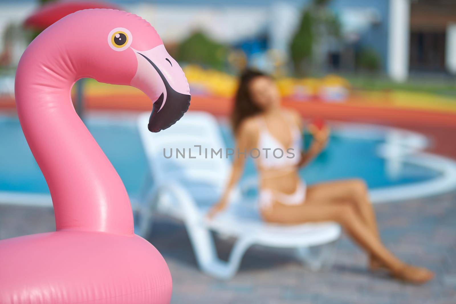 Inflatable pink flamingo ring at swimming pool area outside. Close up view of pink flamingo air mattress with blurred female figure sitting on lounge chair on background. Concept of water activities.