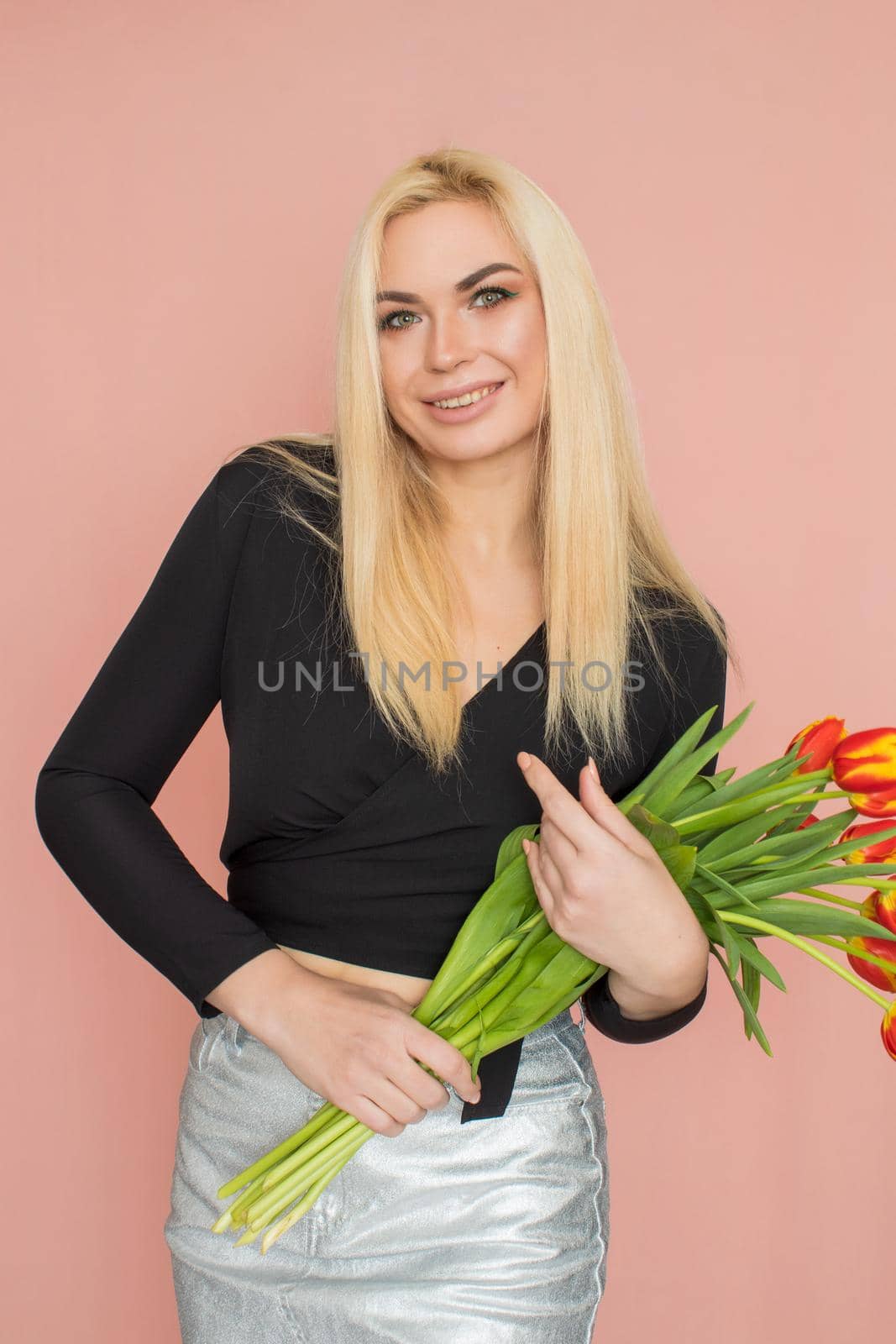 Fashion model woman in fashionable clothes on pink background. Wearing stylish clothing, black blouse, silver skirt. Posing in studio. Holding red tulips in her hands