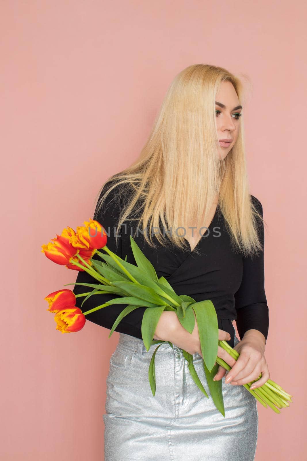 Fashion model woman in fashionable clothes on pink background. Wearing stylish clothing, black blouse, silver skirt. Posing in studio. Holding red tulips in her hands