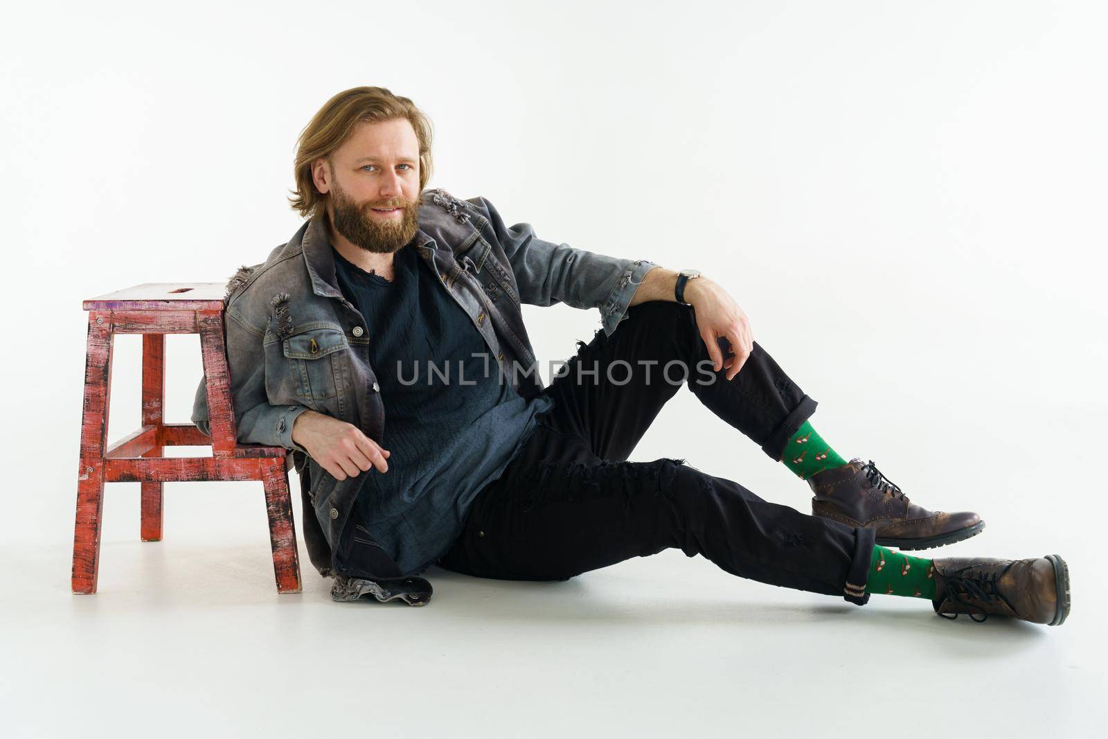 a handsome brutal bearded and long-haired man sits on the floor leaning on a chair on a white background, he is dressed in a denim jacket and pants with bright green socks, dressed like a hipster. High quality photo