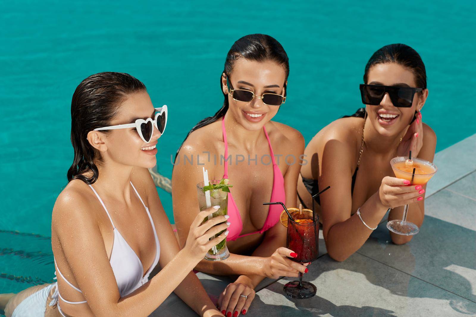 Three glamorous ladies in sunglasses sunbathing, having fun on swimming pool deck in summer. Front view of smiling women in bikinis relaxing, drinking cocktails in blue pool water. Concept of relax.