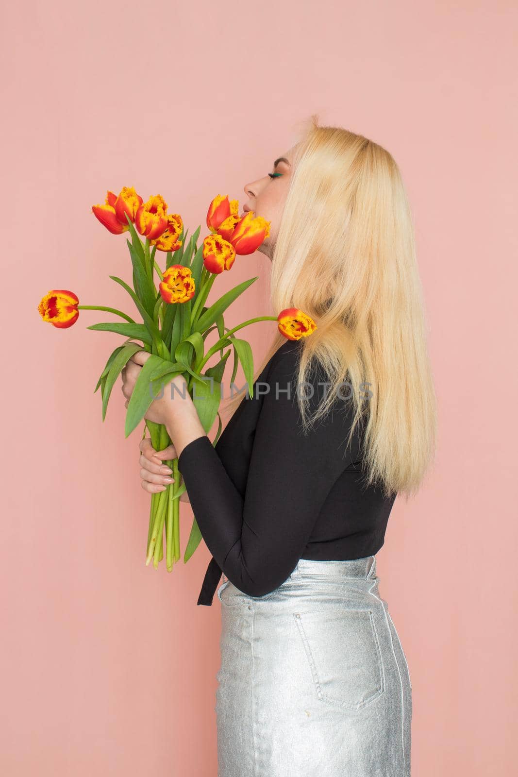 Fashion model woman in fashionable clothes on pink background. Wearing stylish clothing, black blouse, silver skirt. Posing in studio. Holding red tulips in her hands