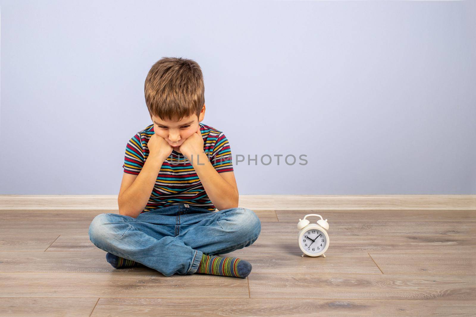 A cute little boy in a T-shirt and jeans is sitting on the floor next to the clock waiting. The boy's pose is tired of waiting.