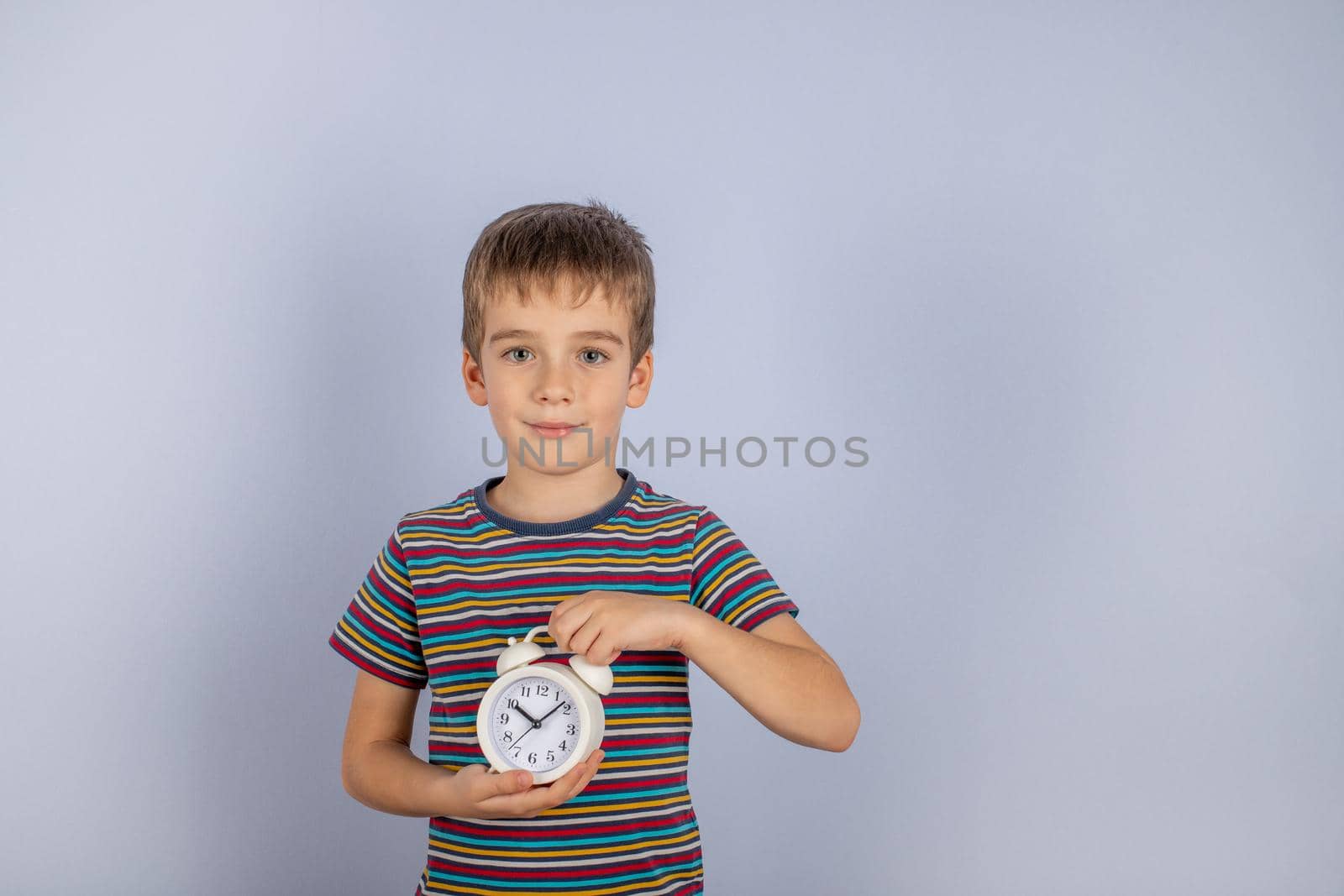 A little boy in a striped T-shirt is holding a white alarm clock. On a blue background. by bySergPo