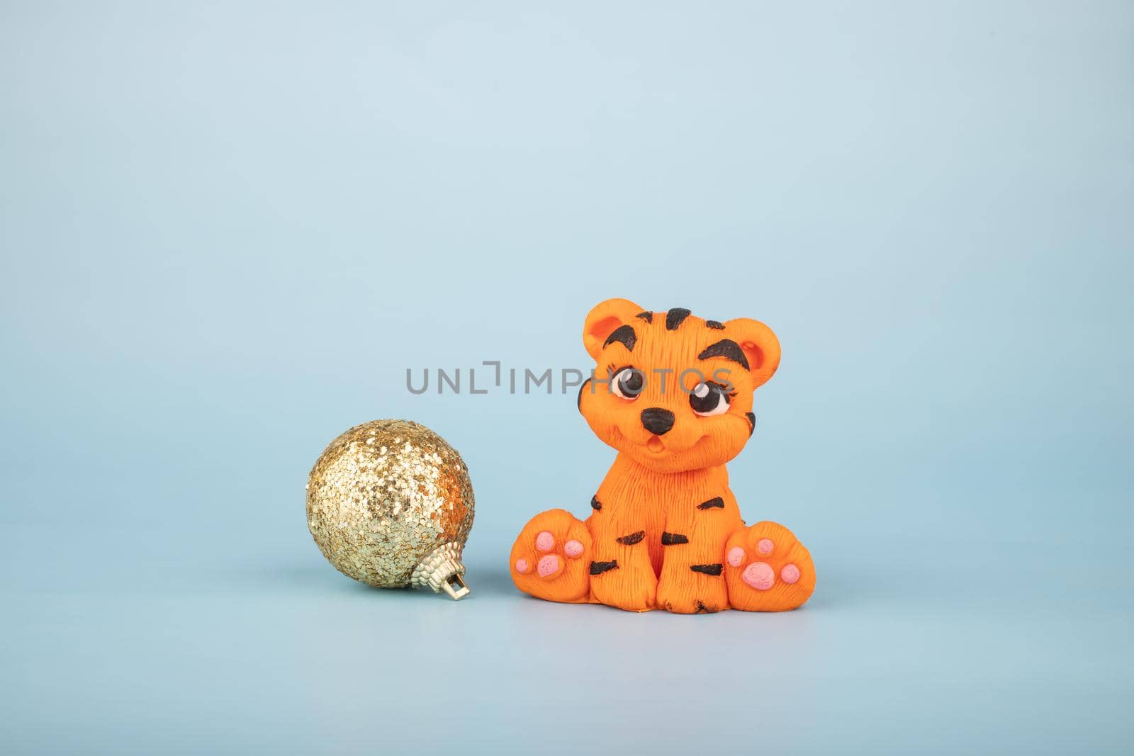 A hand-sculpted orange tiger figurine on a blue background with a golden Christmas tree toy. The year 2022 is the year of the tiger according to the Eastern calendar.