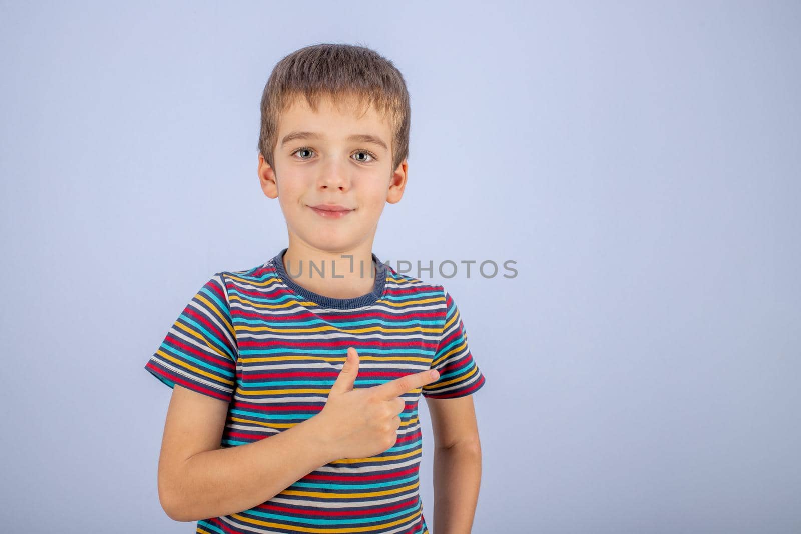 A happy little boy in a striped T-shirt points his finger at the place to copy and smiles on a blue background.