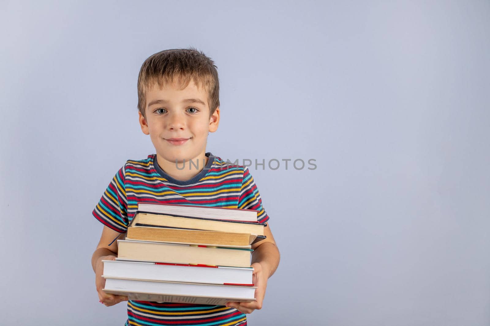 Time To Explore The Concept. A small novice schoolboy holding a stack of books on a blue studio background. Preparation for study. Space for copying.